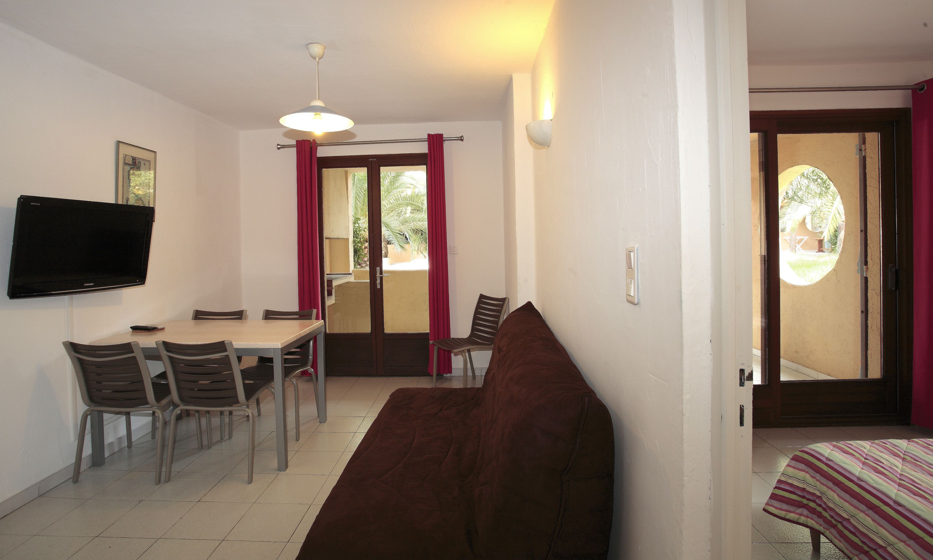 A room with a TV mounted on the wall, a dining table with chairs, a sofa, and a bed with a striped blanket.