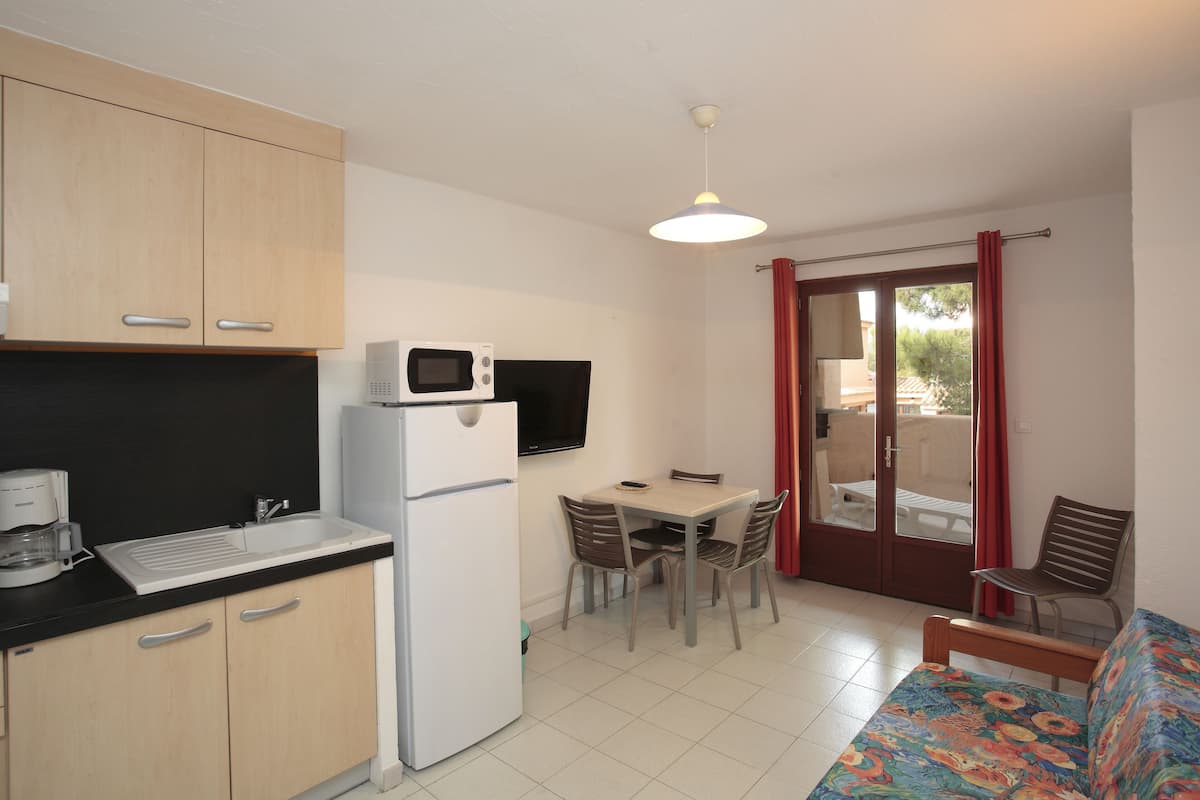A compact kitchen with a microwave, refrigerator, and coffee maker. There is a dining area with a table and chairs, and a sofa with a colorful cover. A sliding glass door leads to an outdoor area.