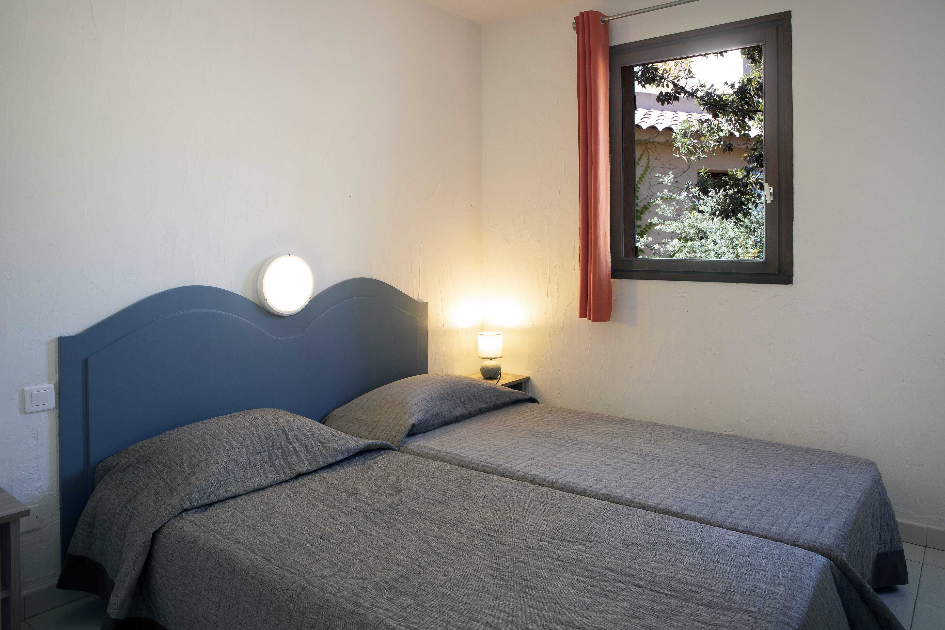 Ein Schlafzimmer mit zwei Betten, einem Nachttisch mit Lampe und einem Fenster mit Blick auf Grünflächen.