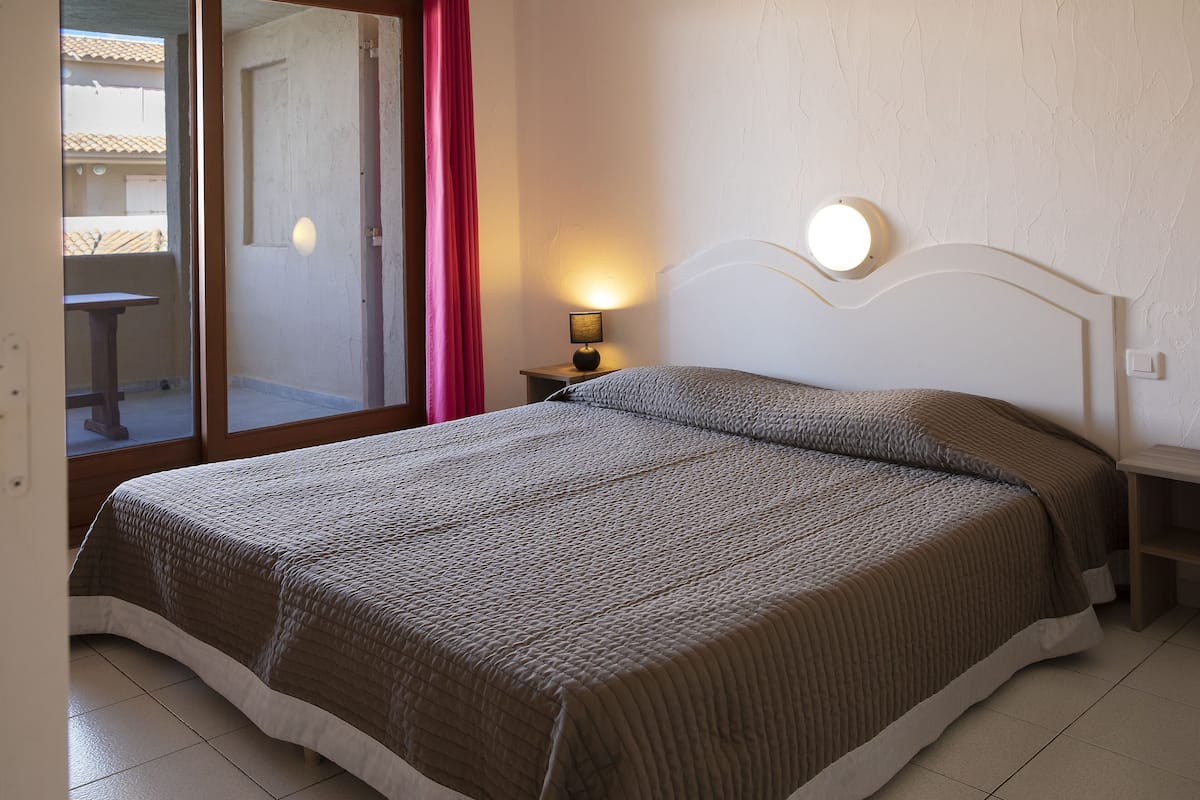 A bedroom with a bed, a nightstand with a lamp, a window with a view of the outside, and a door leading to a balcony.