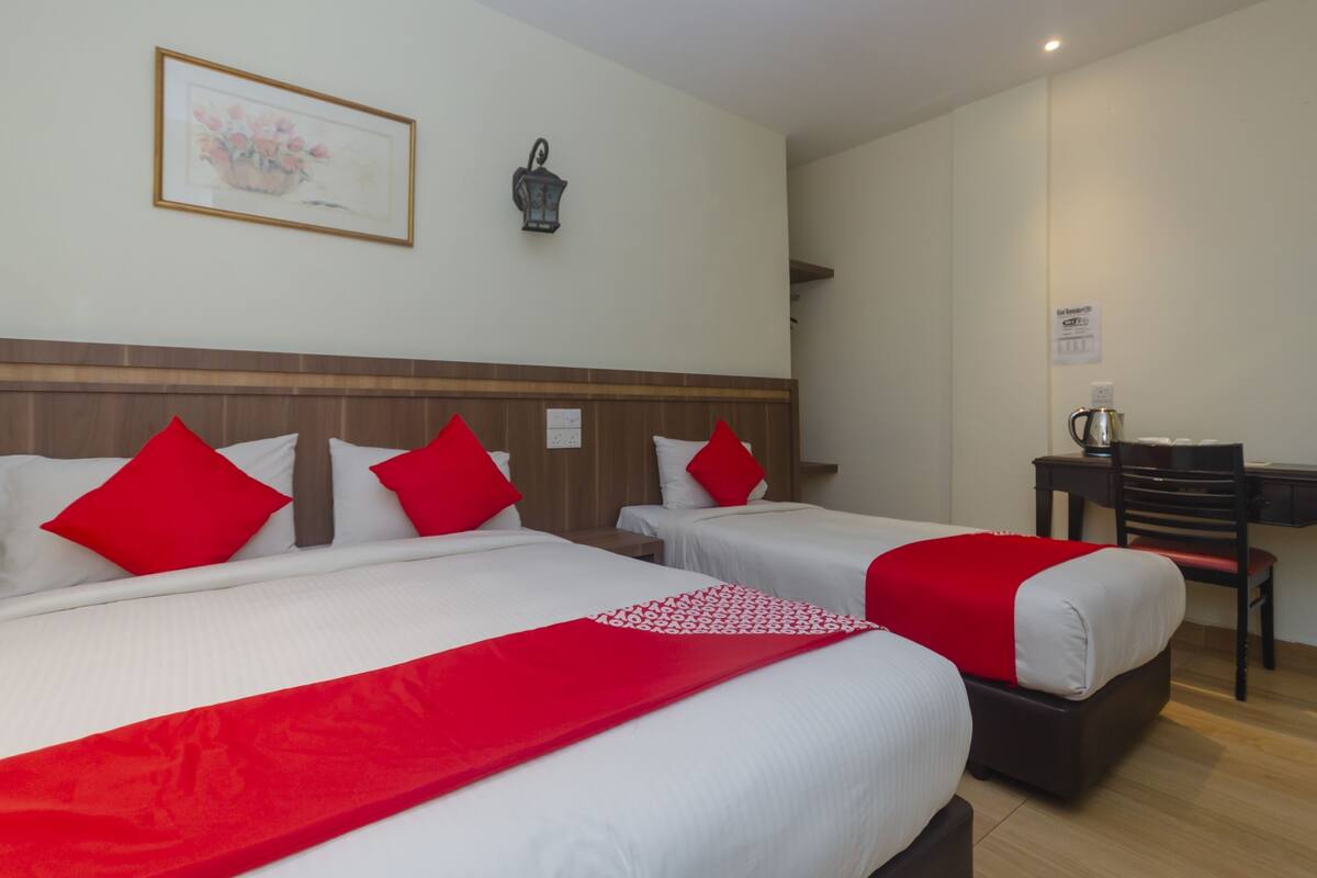 Habitación de hotel con dos camas, cada una con ropa de cama roja y blanca, cabeceros de madera, una mesita con una silla y una lámpara fijada a la pared.