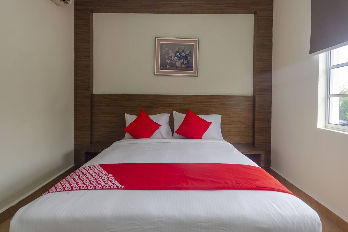Una habitación de hotel con una cama grande, dos almohadas rojas, un cuadro en la pared y una ventana a la derecha.