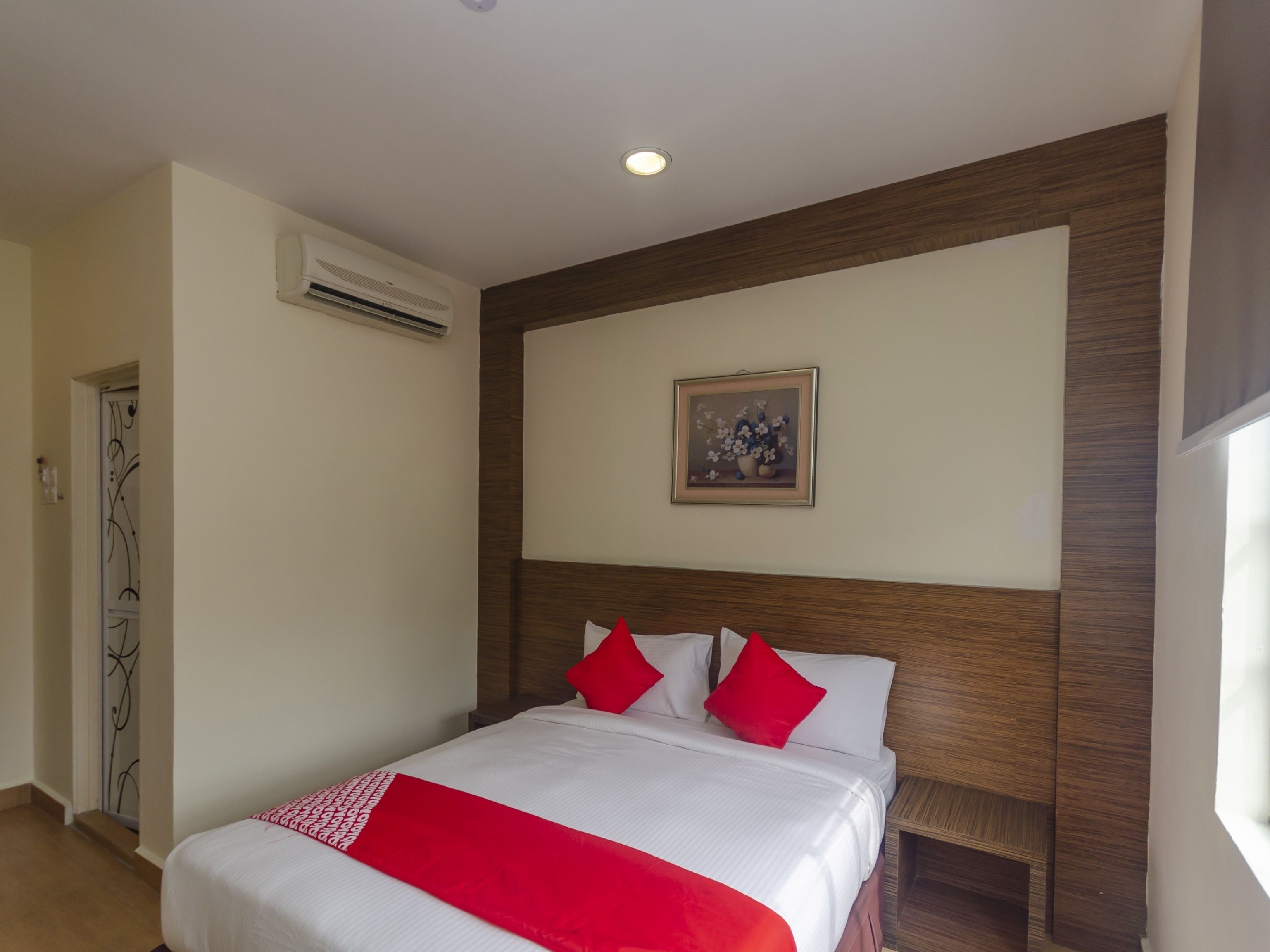 Una habitación de hotel con una cama grande, ropa de cama roja y blanca, un cabecero de madera, un equipo de aire acondicionado y un cuadro en la pared.