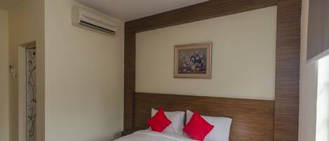 Una habitación de hotel con una cama grande, ropa de cama roja y blanca, un cabecero de madera, un equipo de aire acondicionado y un cuadro en la pared.