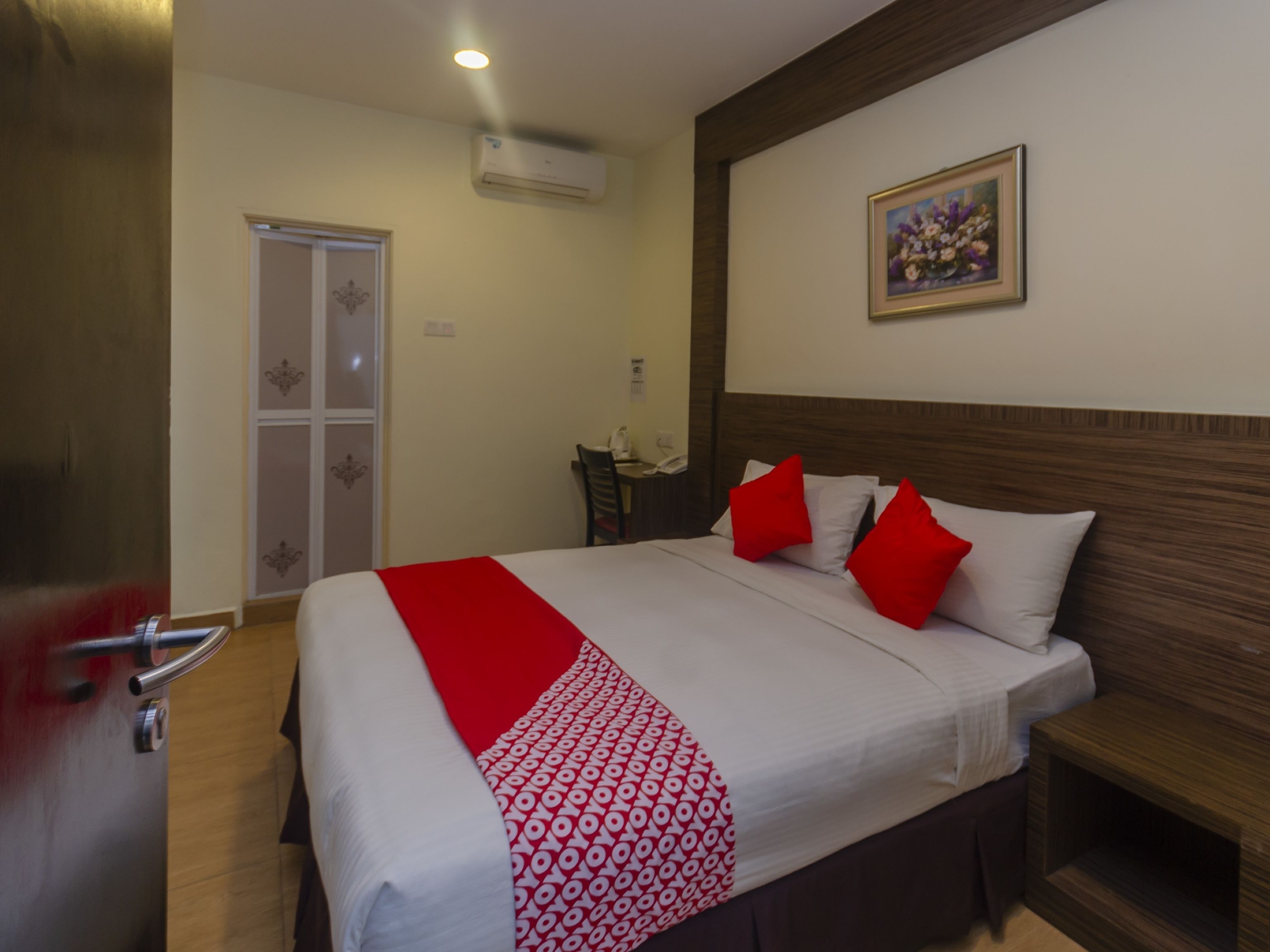 A hotel room with a bed, a bedside table, a chair, a desk, an air conditioning unit, a door, and a painting on the wall.