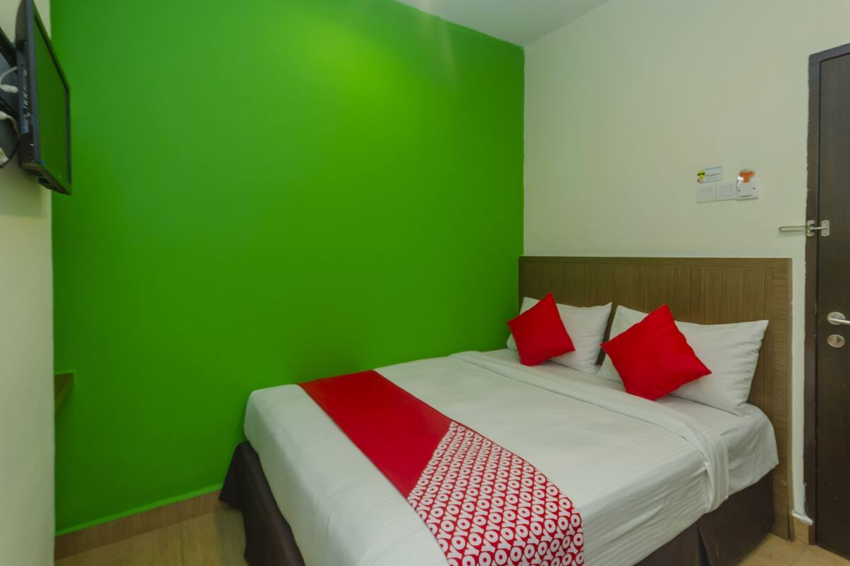Habitación de hotel con pared verde, cama con ropa blanca y almohadas rojas, y cabecera de madera.