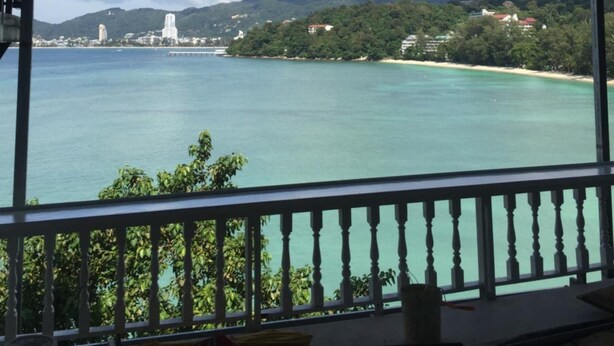 A covered balcony with a view of a turquoise sea, a distant island with buildings, and a cloudy sky. - Nan inn Bungalow (Karon)