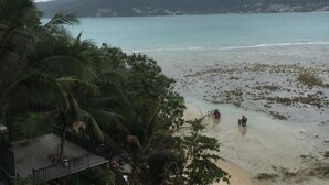 A beach with people walking on the shore, surrounded by palm trees and mountains in the background. - Nan inn Bungalow (Karon)
