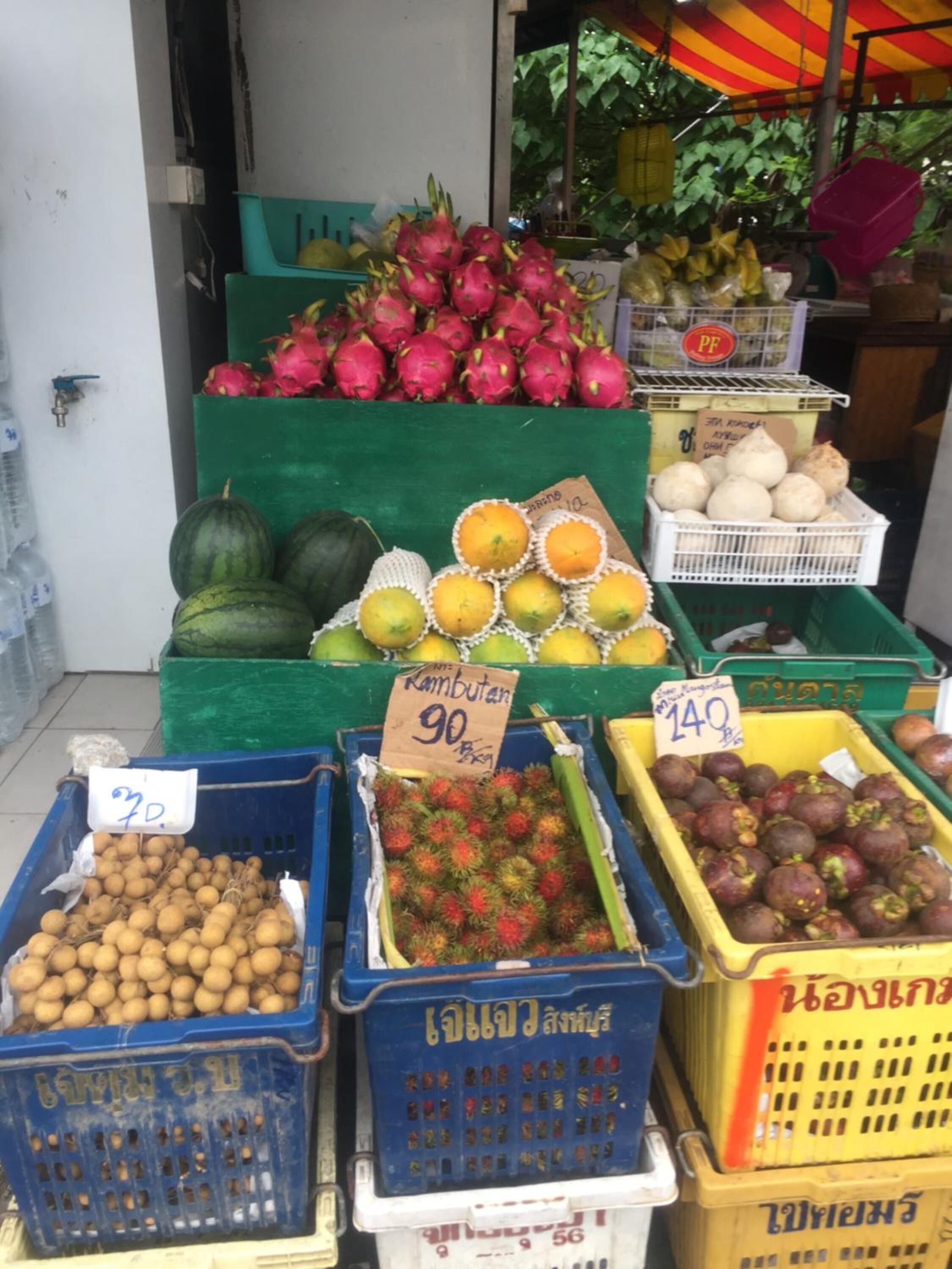 A fruit stand with various fruits like dragon fruit, watermelons, and rambutan. Prices are visible on signs.