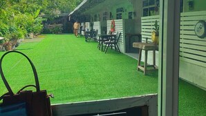 A view of a room with a window overlooking a green lawn and a building with a sloped roof. - Nan inn Bungalow (Karon)