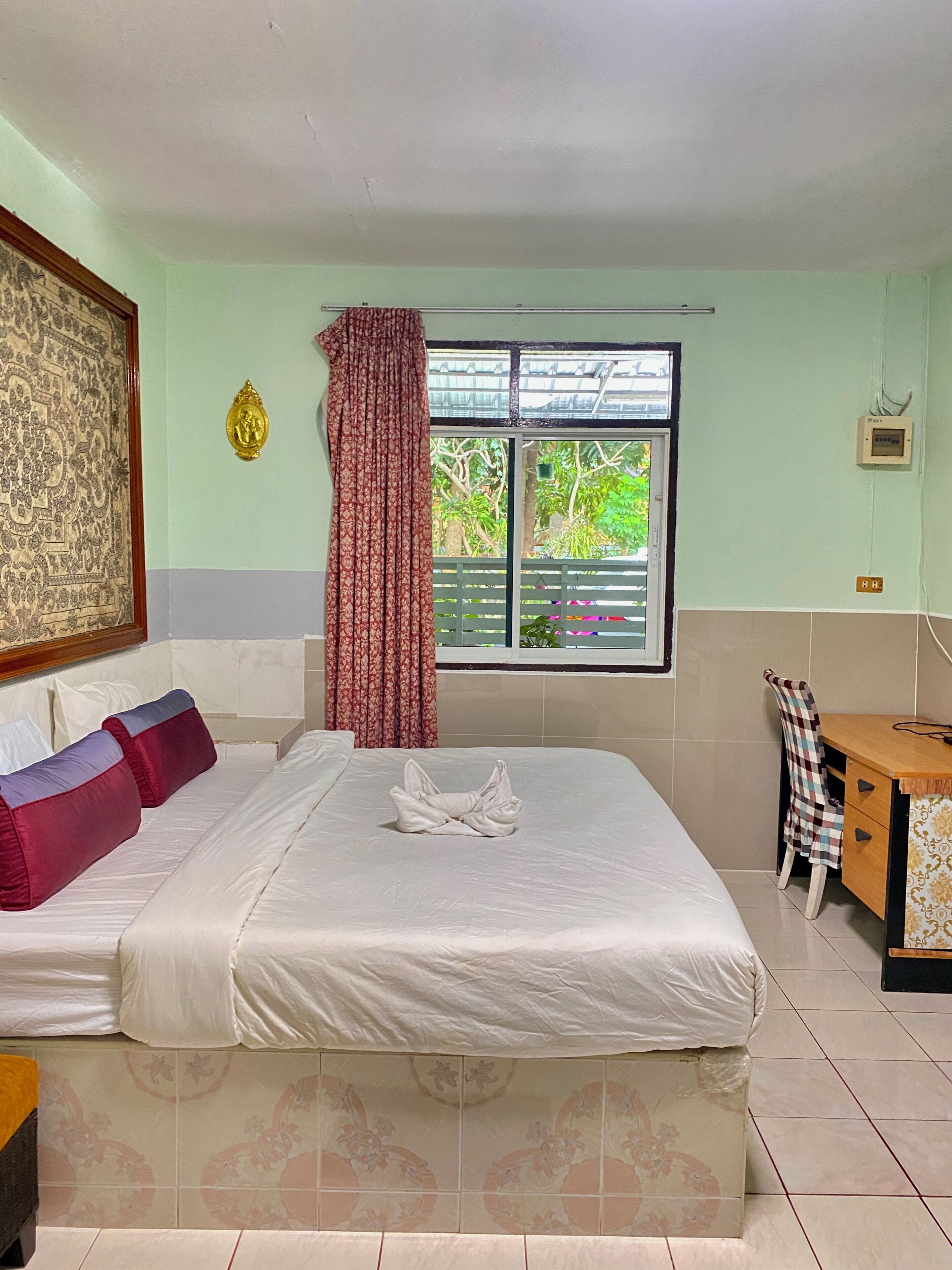 A hotel room with a bed, a desk, a chair, a window with a view of greenery, and a decorative wall hanging.