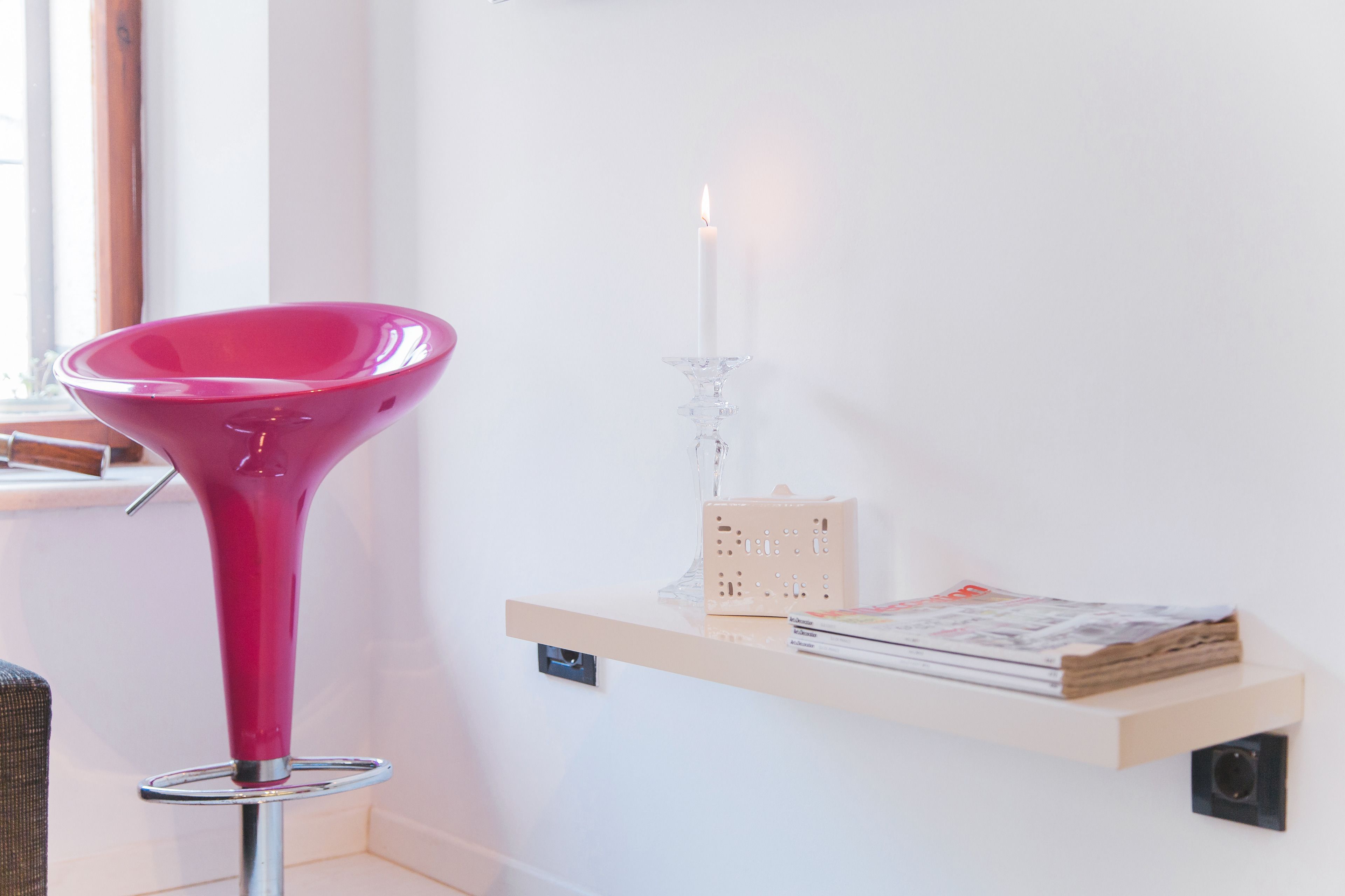 Un interior moderno con una silla cónica rosa, una vela blanca sobre un portalápices de vidrio y un estante de madera con libros y una cajita.