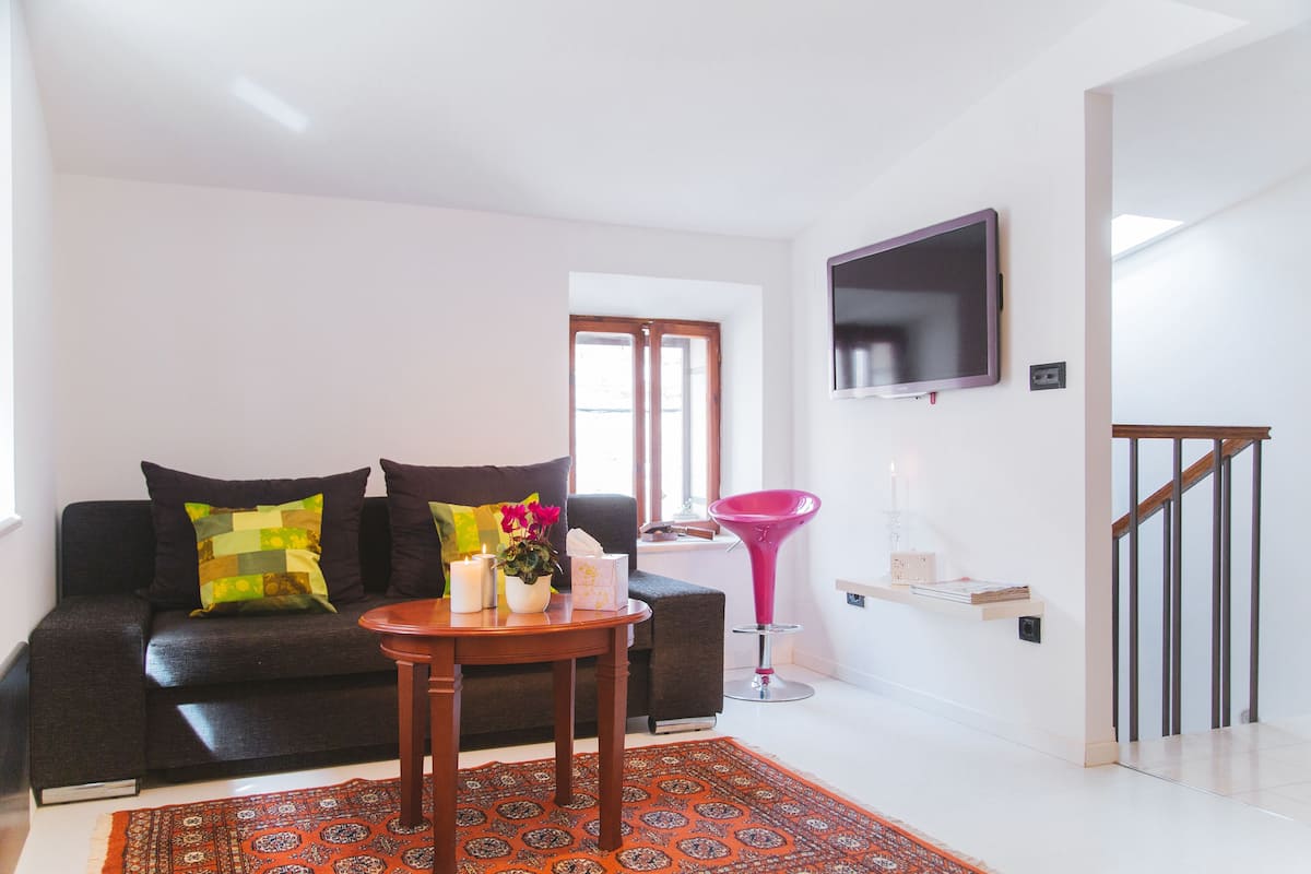 A modern living room with a dark sofa, a small wooden table, a vibrant rug, and a pink chair.