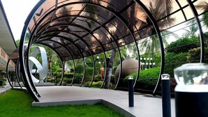 Una estructura arquitectónica moderna con estructura metálica curva y paneles de vidrio, rodeada de follaje y césped bien cuidado.