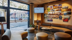 A modern hotel lobby with a large TV screen displaying the hotel name, comfortable seating areas, and a city view through large windows.