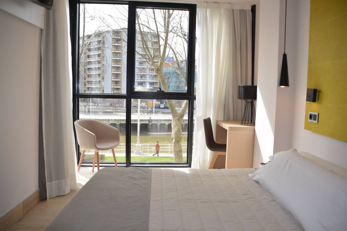 Hotellihuone, jossa on suuri sänky, työpöytä, tuoli ja suuri ikkuna, josta avautuu näkymä kaupungin ylle.