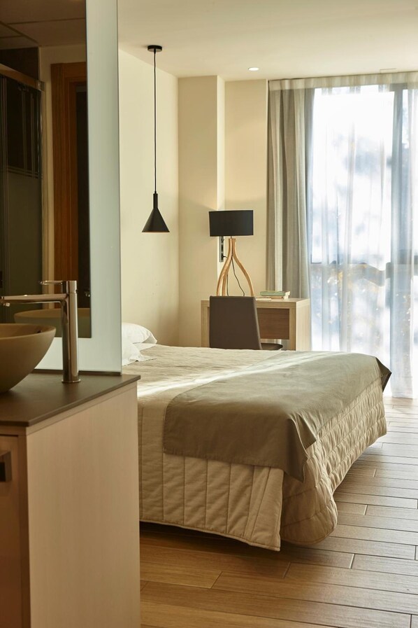 Una habitación de hotel con cama, mesita de noche, lámpara y un amplio ventanal con cortinas.
