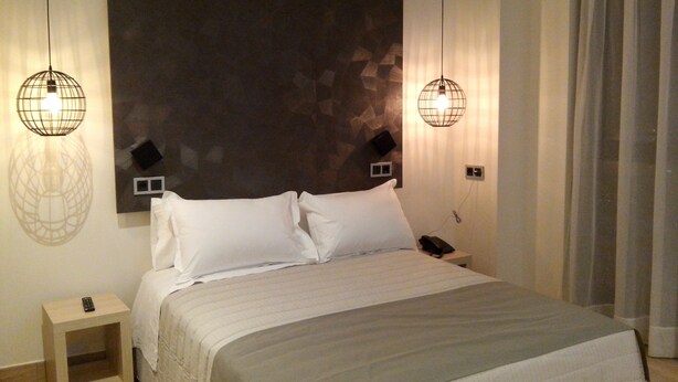 A neatly made bed with white linens, a bedside table with a remote control, and two wall-mounted lamps.