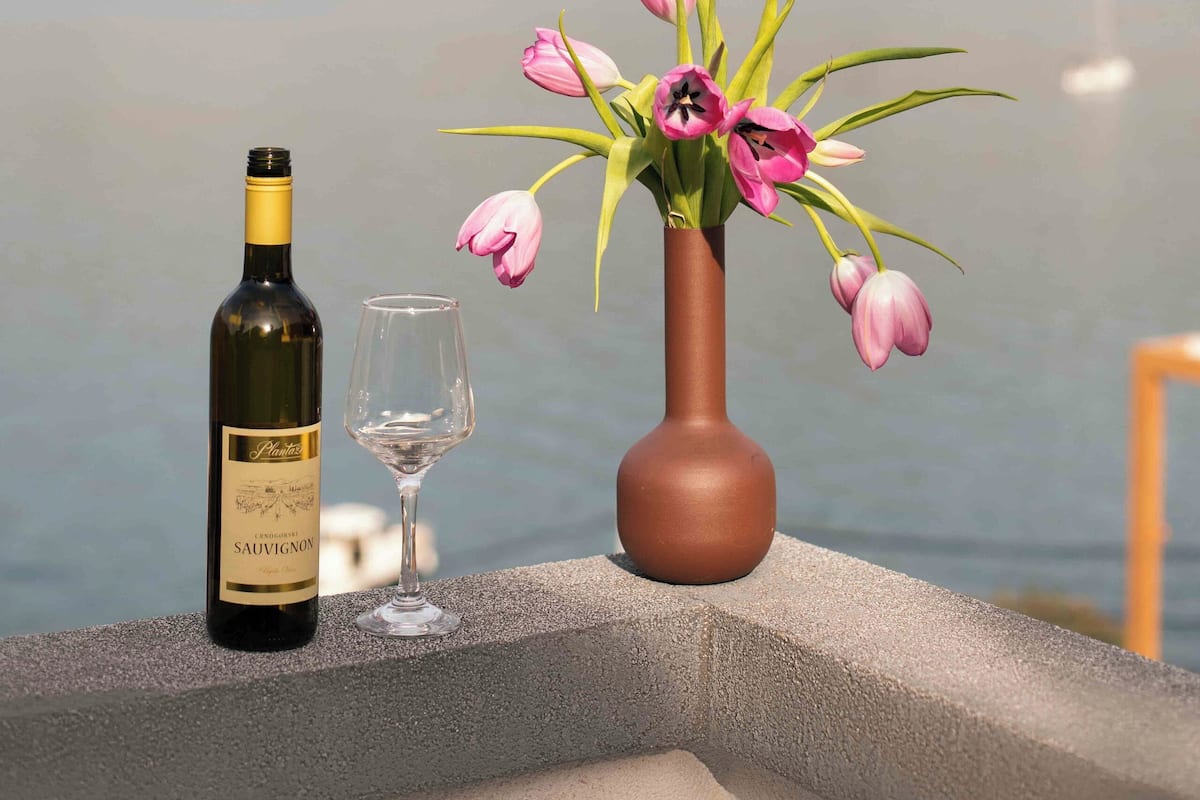 A bottle of wine and a glass on a ledge with a vase of pink flowers and a lake view in the background.