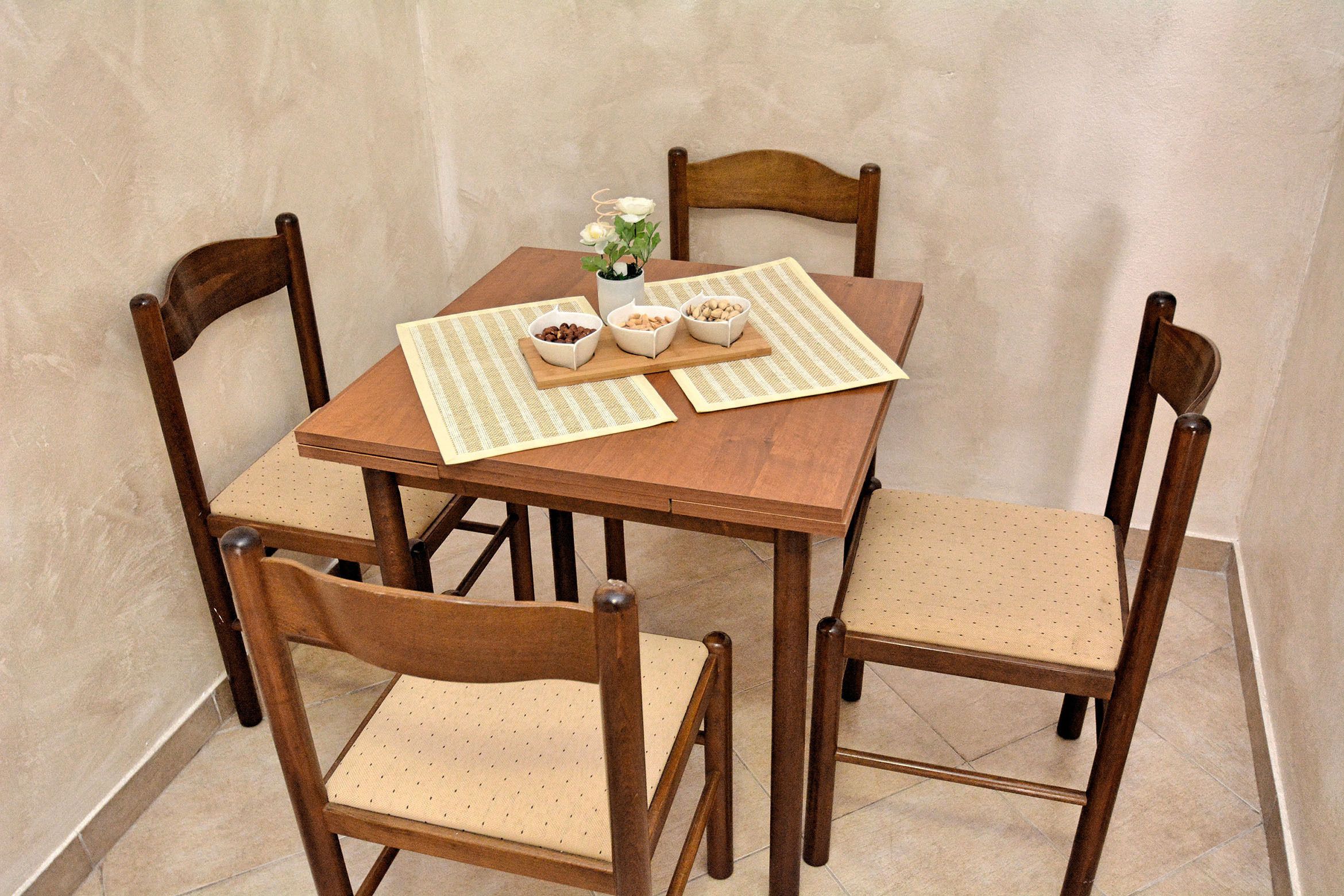 A wooden dining table with four chairs, a vase with flowers, and three bowls of nuts.