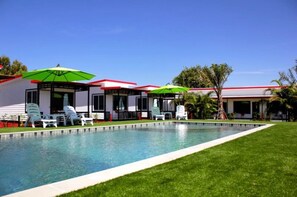 A pool area with lounge chairs and umbrellas, surrounded by a well-maintained lawn and a building with a red roof. - Cape Go Resort (Tha Mai)