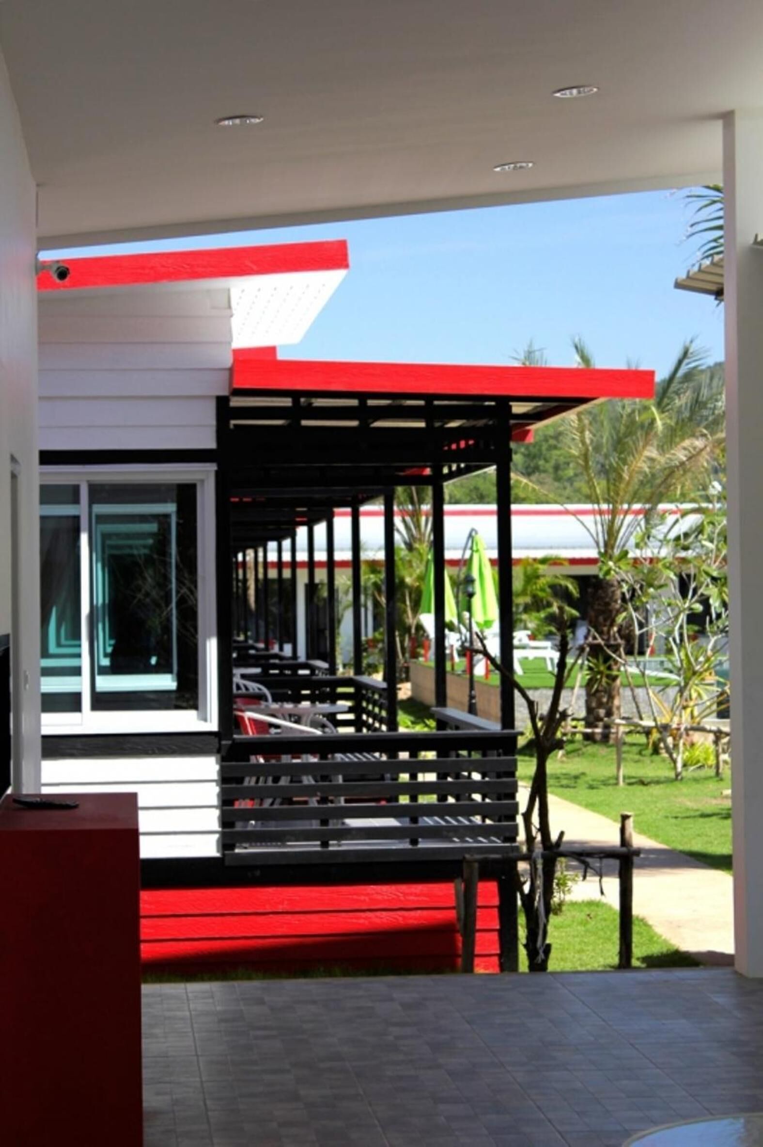 A modern building with a red and white facade, a covered outdoor seating area with black benches, and a view of a green landscape with palm trees.
