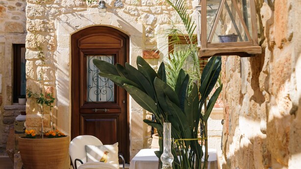A stone building with a wooden door and windows, outdoor seating with tables and chairs, and potted plants. - Casa Di Veneto (Hersonissos)