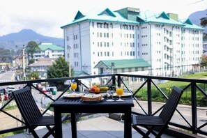 A balcony with a table set for breakfast, overlooking a large building with green roofs. - Cottage 42 (Nuwara Eliya)