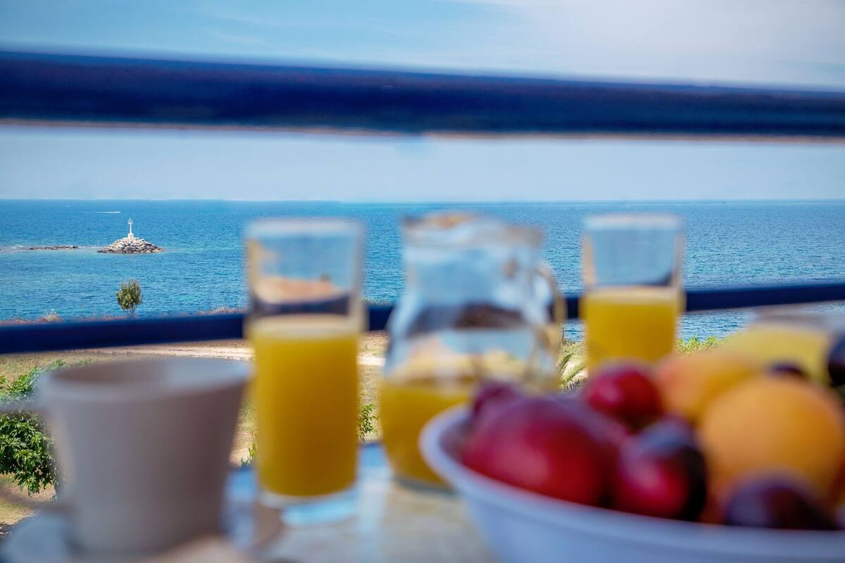 A breakfast table with orange juice, a bowl of fruit, and a view of the sea and a lighthouse.