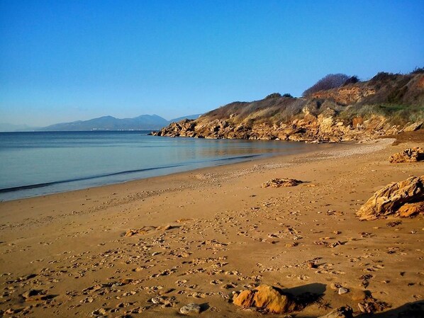 Una playa con aguas claras, costa rocosa y montañas a lo lejos.