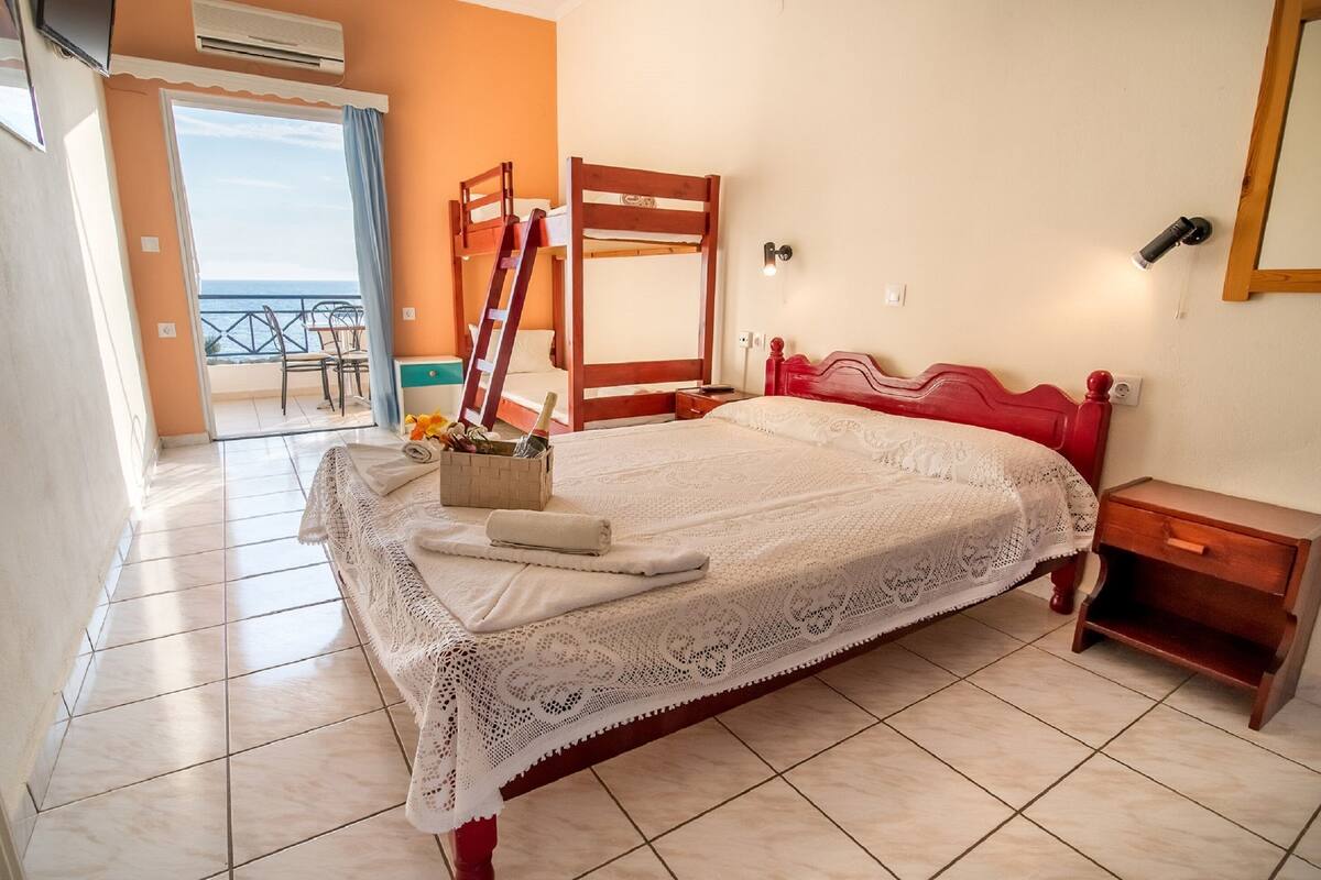A bedroom with a bed, a wooden nightstand, a red headboard, a white lace bedspread, a wooden ladder, and a view of the outside balcony.