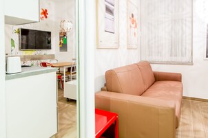 Uma sala de estar moderna com um sofá bege, uma mesa lateral vermelha e uma área de cozinha com micro-ondas e mesa de jantar.