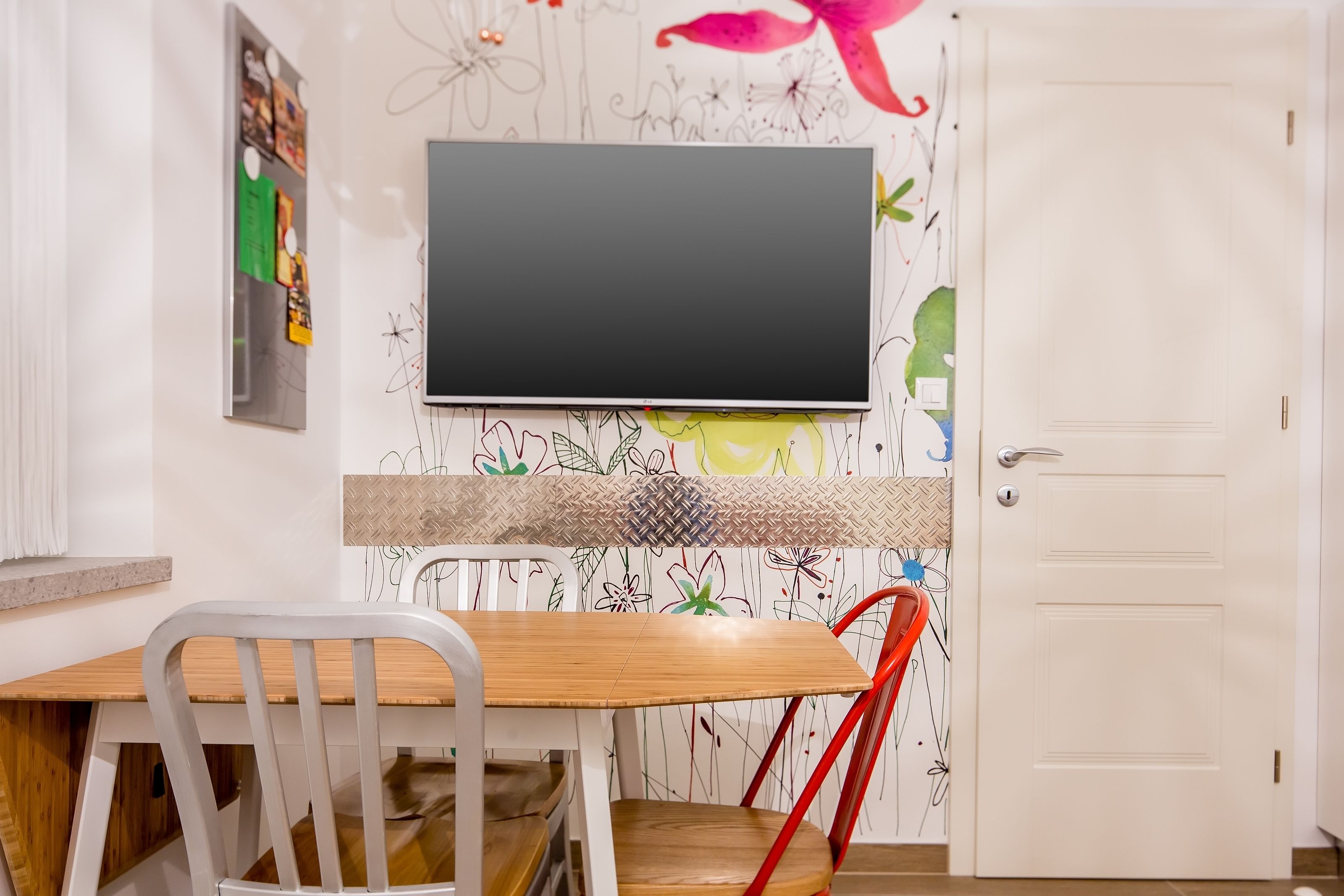 A room with a wooden table, white chairs, a flat-screen TV mounted on the wall, and a white door.