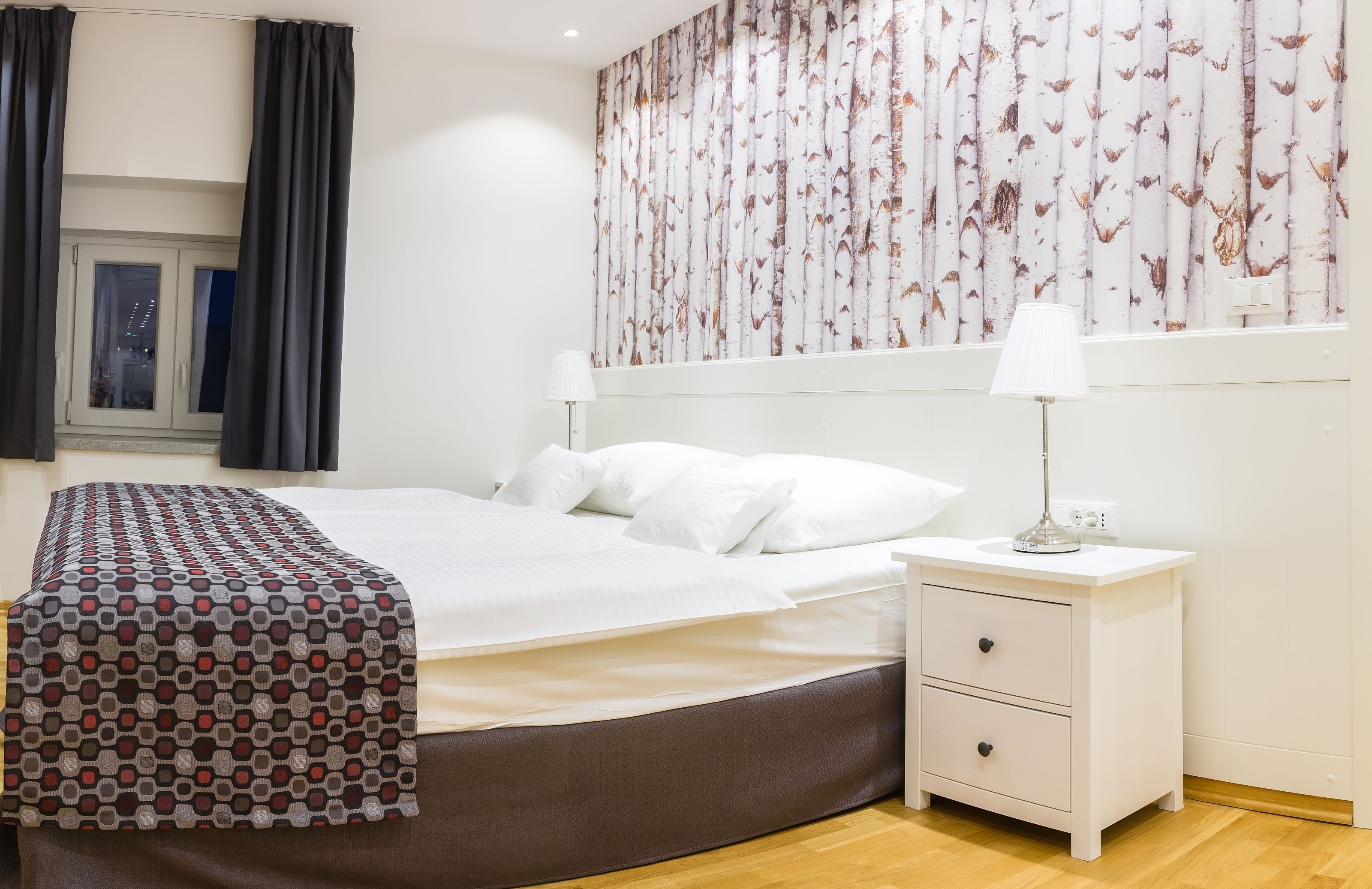 A bedroom with a bed, a nightstand, a lamp, a window with curtains, and a wall with a birch tree wallpaper.