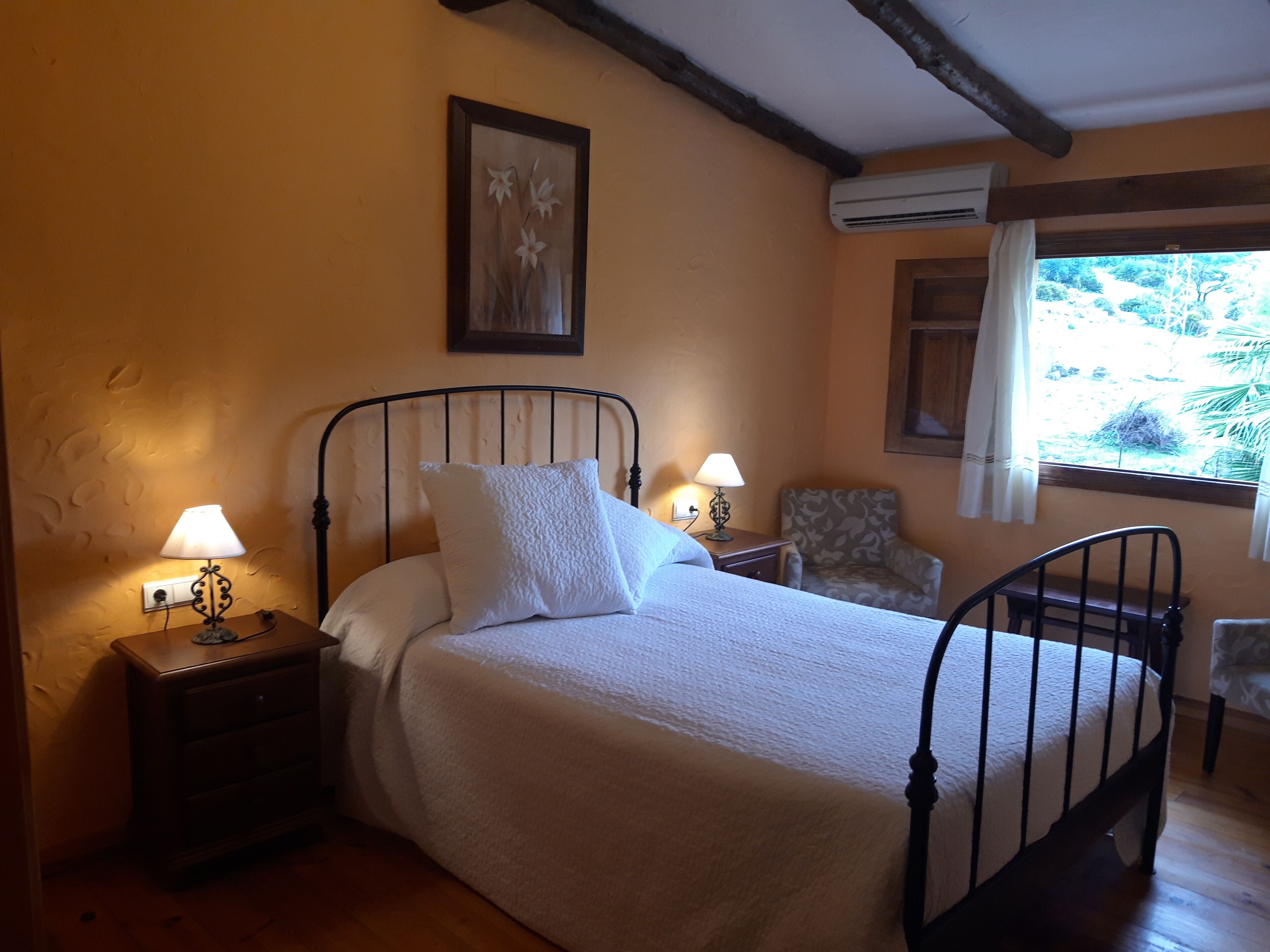 Ein Schlafzimmer mit einem Bett, zwei Nachttischen mit Lampen, einem Sessel, einem Fenster mit Vorhängen und einer Klimaanlage.