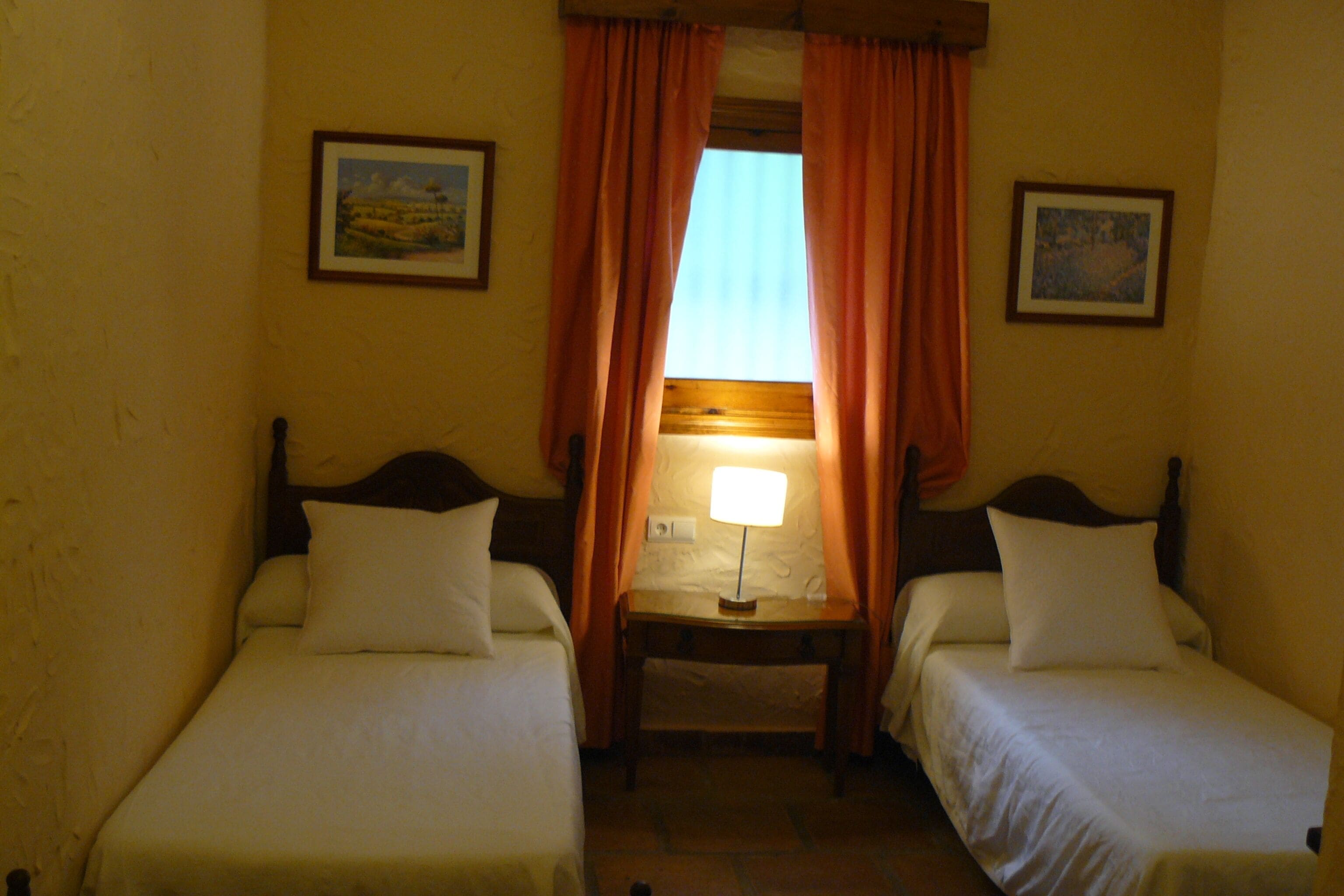 Una habitación con dos camas individuales, una mesita con una lámpara y dos cuadros en las paredes.