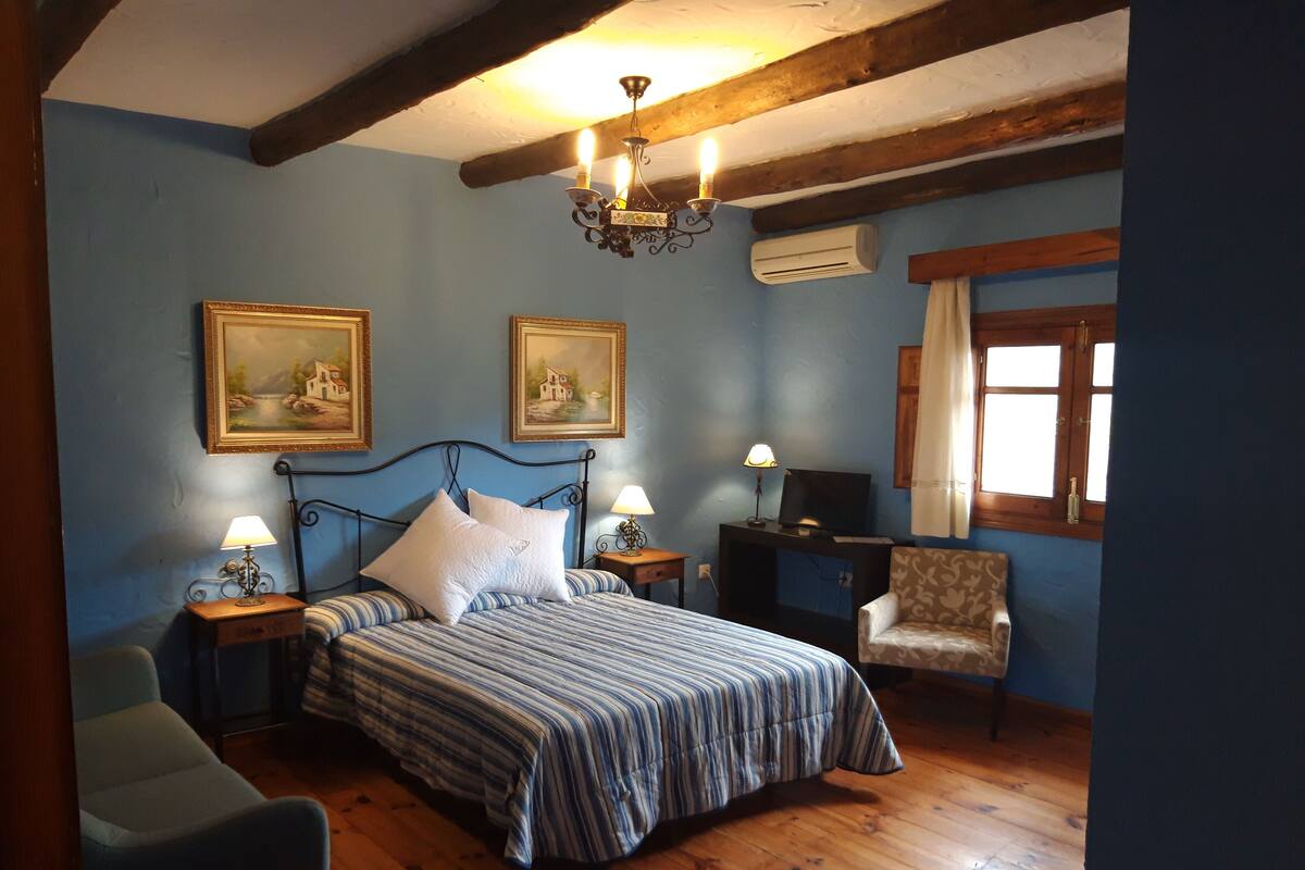 Ein Schlafzimmer mit einem Bett, zwei Nachttischen mit Lampen, einem Sessel, einem Schreibtisch mit Computer und zwei Bildern an der Wand.