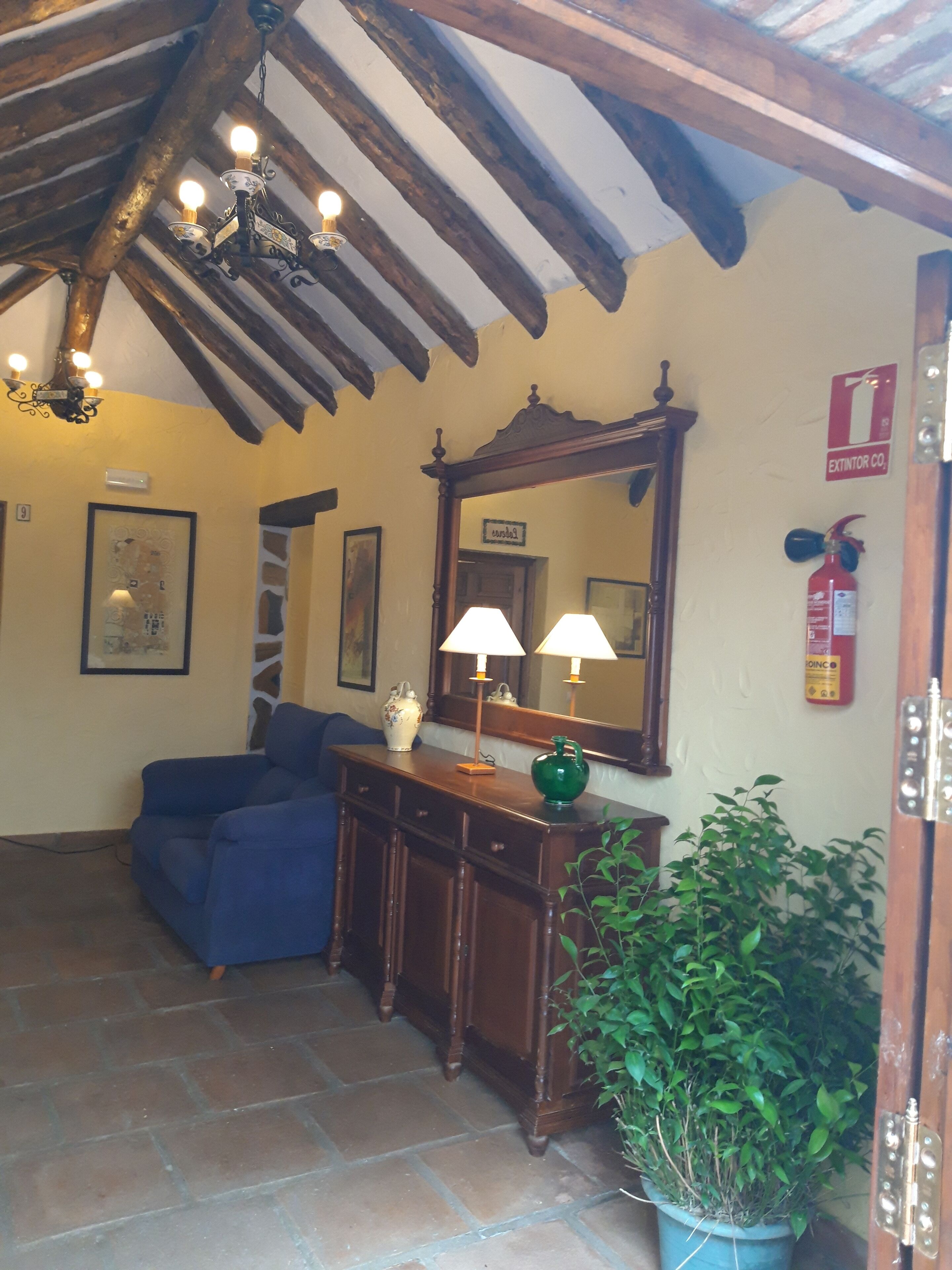 A room with a wooden ceiling, a blue sofa, a wooden console table with a green vase, a mirror, and a fire extinguisher.