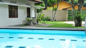 Une piscine à l’eau d’un bleu limpide, entourée d’un bâtiment blanc doté d’un toit en tuiles, d’un petit jardin et d’une clôture verte.