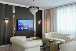 A modern living room with a large flat-screen TV, two white leather sofas, a round gold table, and a crystal chandelier. - Aleksander Palace (Skopje)