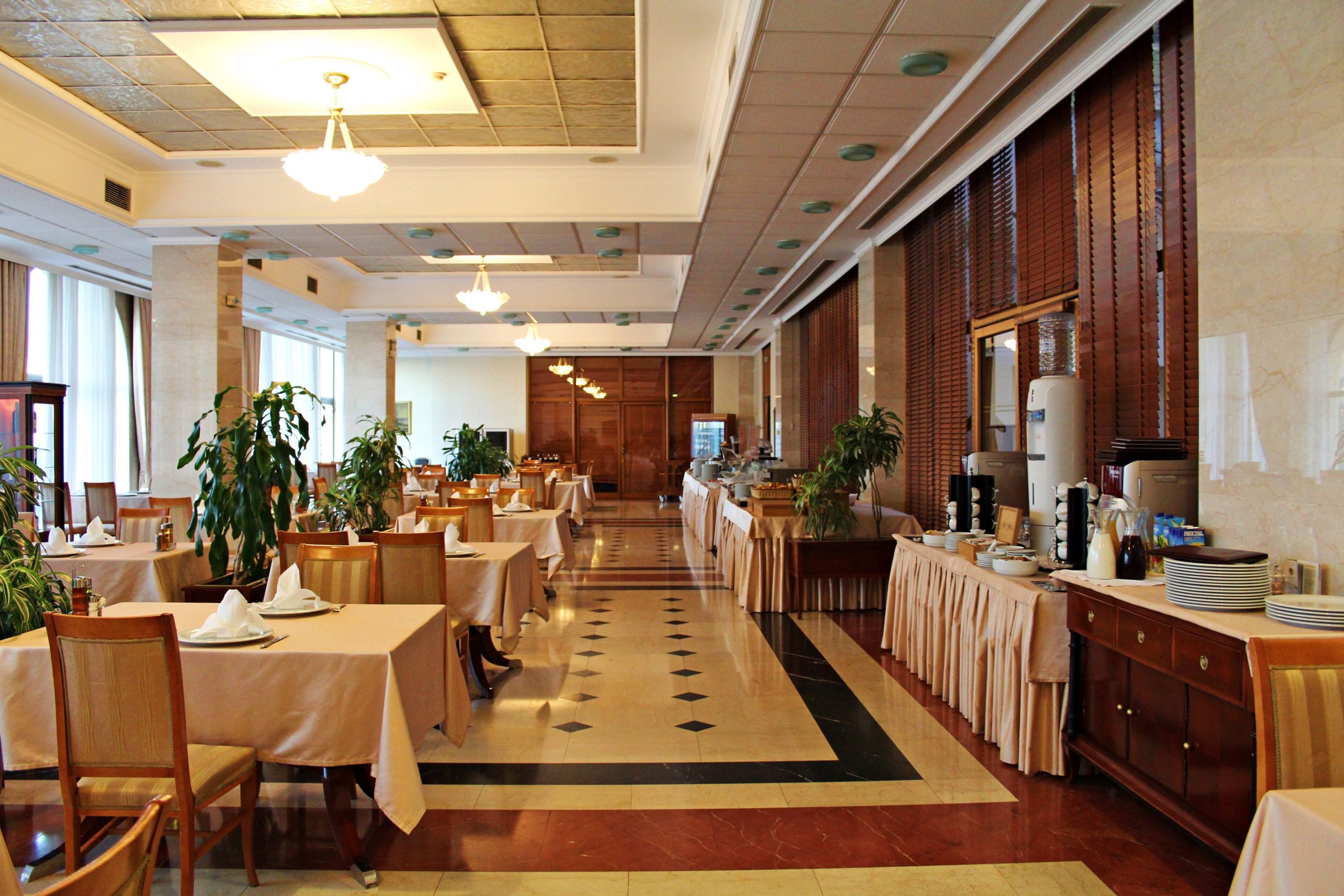 A spacious hotel dining area with multiple tables and chairs, a central aisle, and a buffet setup on the right.