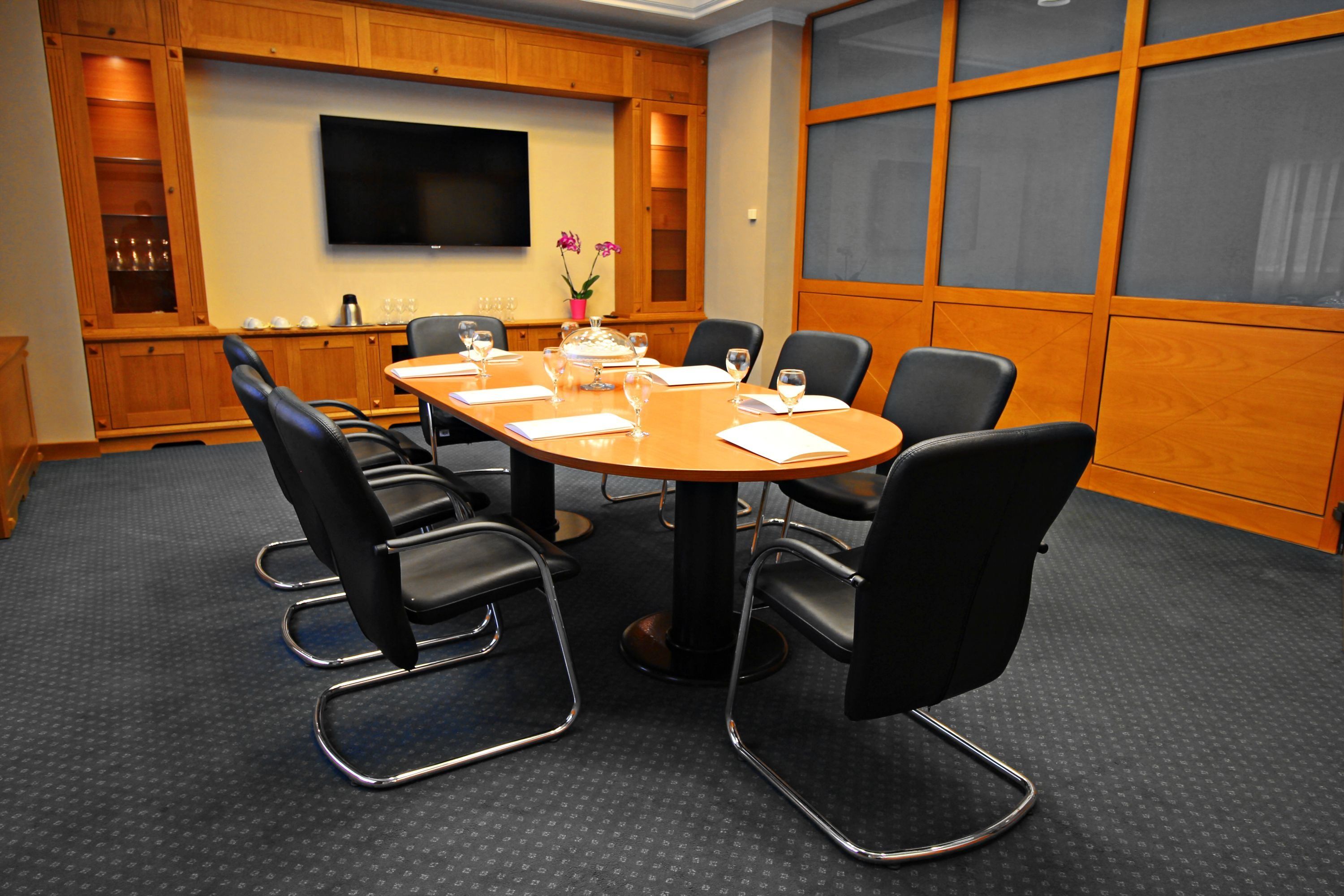 A conference room with a round table, black chairs, a flat-screen TV, wooden cabinets, and a small potted plant.