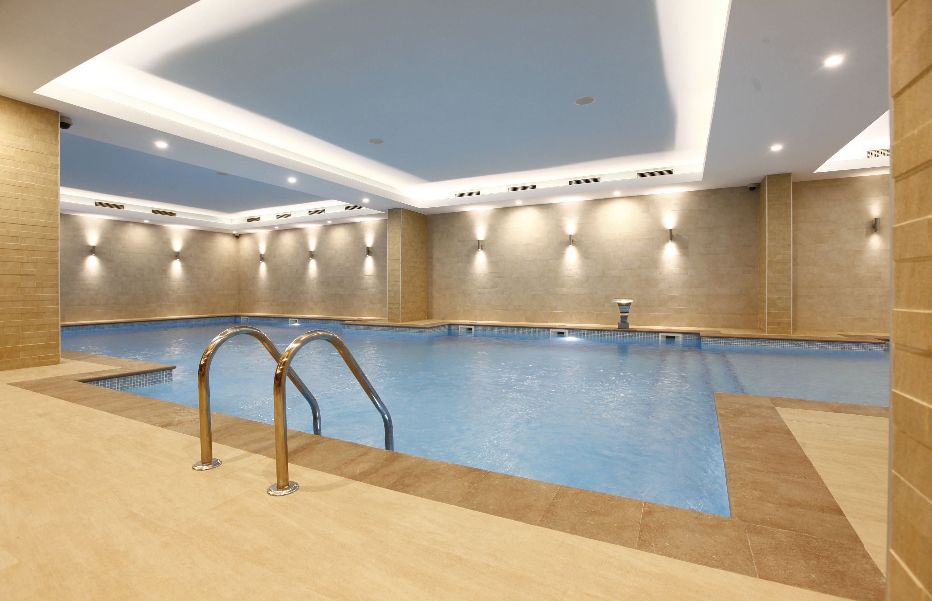 An indoor swimming pool with a blue water feature, surrounded by beige walls and a tiled floor.
