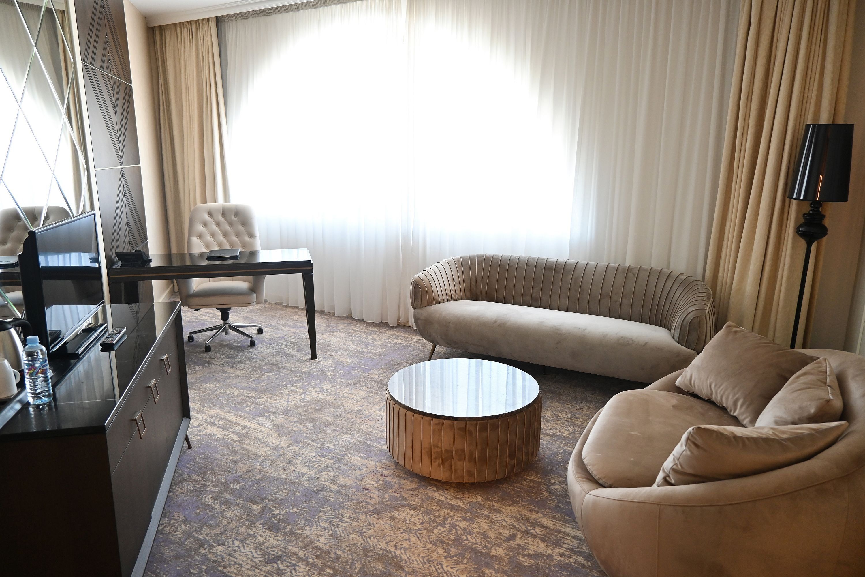 Habitación de hotel con sofá, sillón, mesa de centro redonda y escritorio con computadora.