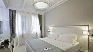 Quarto de hotel com cama, luminárias laterais, televisão e secretária com candeeiro.