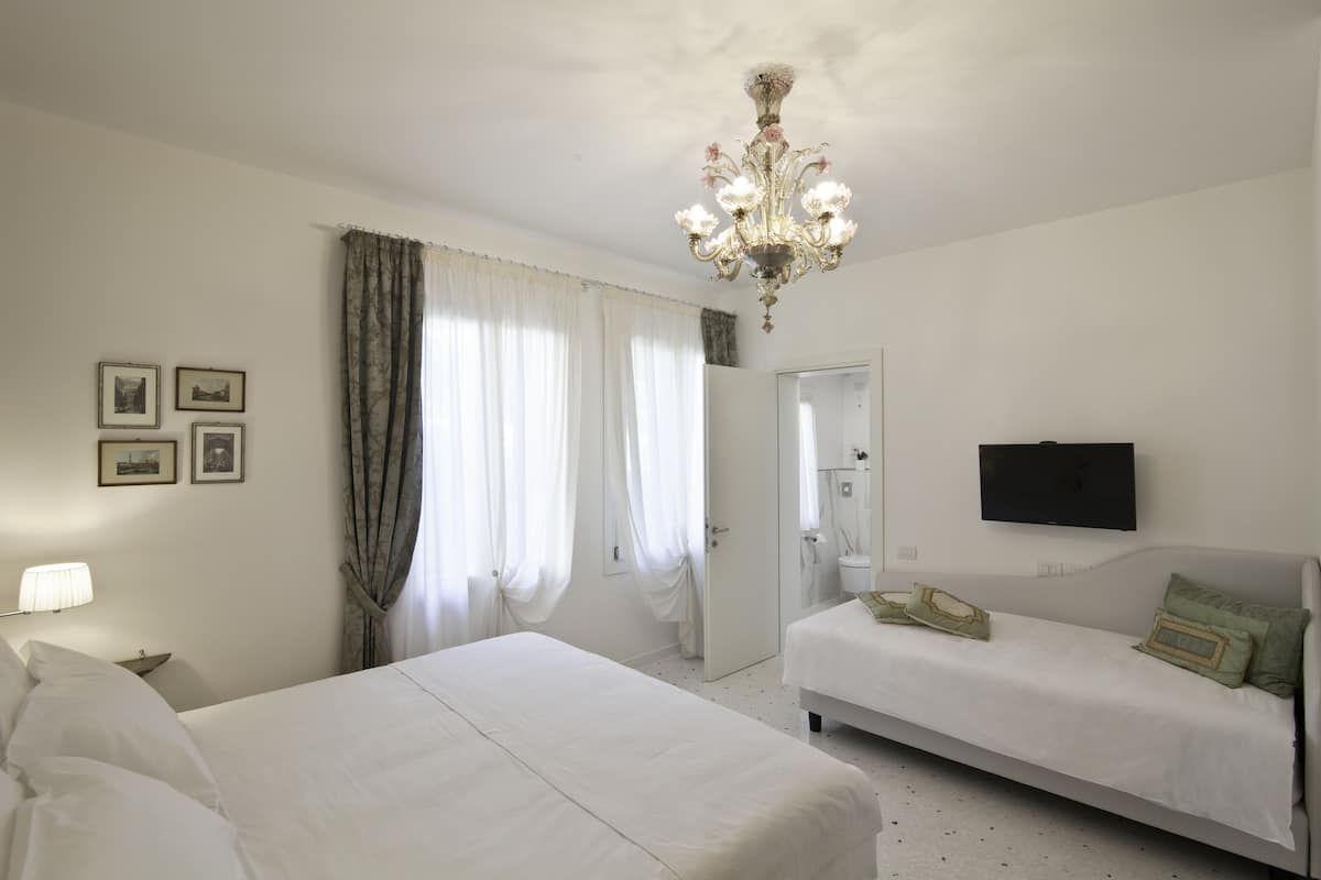 A hotel room with a large bed, a smaller bed, a TV mounted on the wall, and a chandelier.