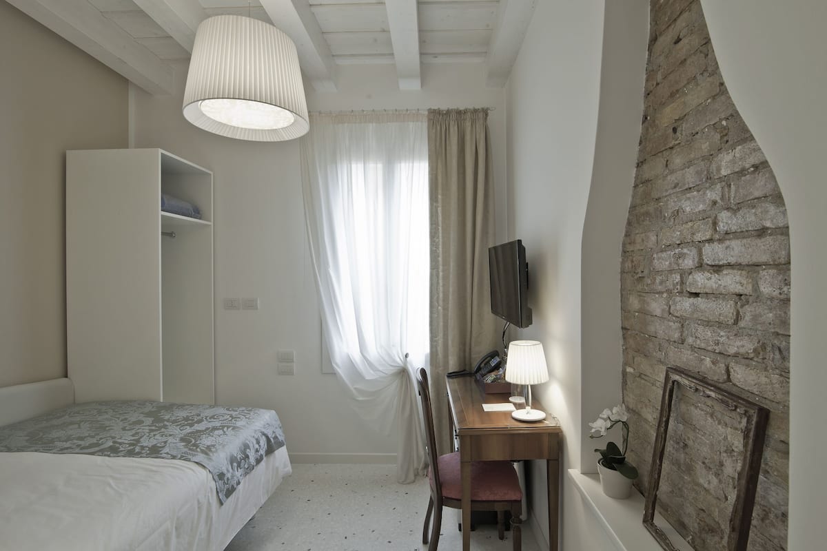 A bedroom with a bed, a desk with a lamp, a wardrobe, a television, and a brick wall.