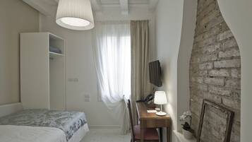 A bedroom with a bed, a desk with a lamp, a wardrobe, a television, and a brick wall.