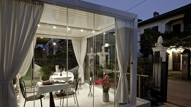 Uma área de refeições envidraçada com mesa e cadeiras, um vaso com flores e vista para a rua.