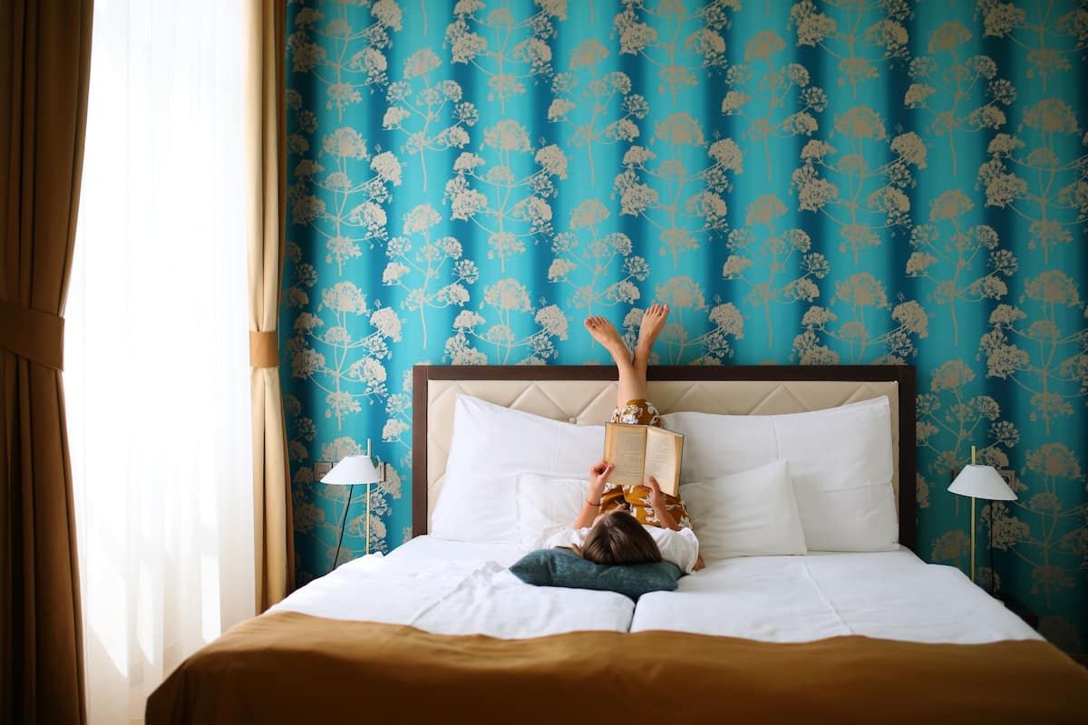 Una cama con cabecera blanca, una persona acostada boca arriba leyendo un libro, y una pared con estampado floral al fondo.